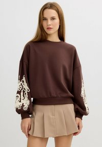 Sudadera marrón con mangas oversize que presenta bordados de flores blancas. Textura suave, cuello redondo y dobladillo acanalado, combinada con una falda beige.