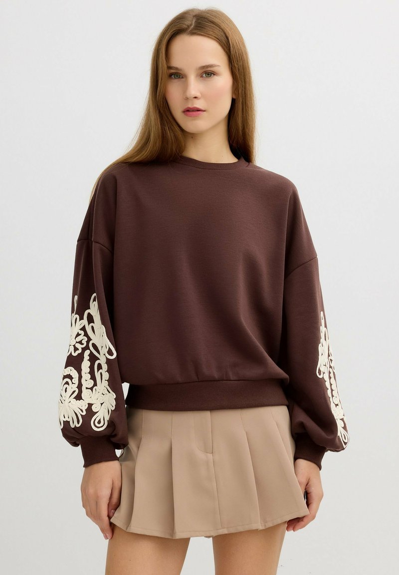 Sudadera marrón con mangas oversize que presenta bordados de flores blancas. Textura suave, cuello redondo y dobladillo acanalado, combinada con una falda beige.