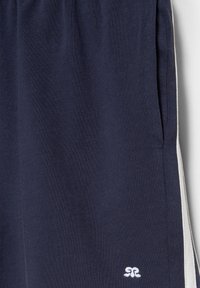 Tissu bleu marine avec poche latérale, bande verticale blanche le long du bord et petit logo blanc brodé près du bas.