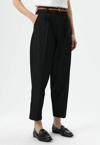 Zwarte, hooggetailleerde, geplooide broek van gestructureerde stof met zijzakken, gecombineerd met een gevlochten bruine riem en zwarte loafers.