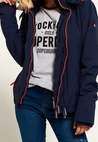 Marine puffer jakke med kontrasterende røde lynlåse, lynlåslommer og høj krave. Sat sammen med en grå grafisk t-shirt og blå jeans.
