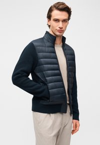 Μπουφάν σε navy από φουσκωτό υλικό με πλεκτά μανίκια με υφή. Διαθέτει ψηλό γιακά, κλείσιμο με φερμουάρ και εφαρμοστή σχεδίαση. Φορώντας το πάνω από ελαφρύ μπεζ πουκάμισο.
