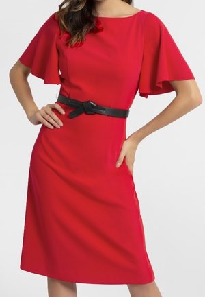 Frau in einem knielangen roten Kleid mit flatternden Ärmeln und schwarzem Gürtel, stehend mit den Händen in die Hüften gestützt vor weißem Hintergrund.