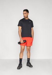 Μαύρο αθλητικό μπλουζάκι, κόκκινα σορτς, μαύρες κάλτσες και μαύρα αθλητικά παπούτσια. Κρατάει μια μαύρη παγούρι νερού, στέκεται μπροστά σε απλό φόντο.