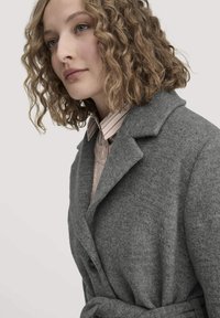 Grauer Wollmantel mit Reverskragen, doppelreihiger Knopfleiste und tailliertem Gürtel. Darunter sind ein hellrosa Pullover und ein gestreiftes Hemd sichtbar.