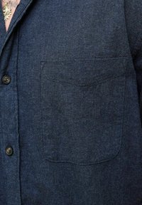 Navy blue textured shirt with front pocket, black buttons, and a gold sun-shaped pendant necklace partially visible at collar.