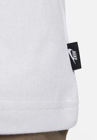 Närbild av vit tygssöm med svart Nike-logotagg fäst på sidan ovanför kläder i tan-färg.