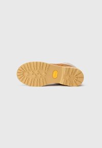 Sål af en tan vinterstøvle med dybe slidbaner og et gult Vibram-logo, vist mod en ensfarvet lys baggrund.