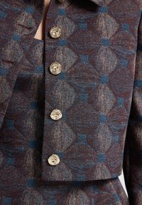 Chaqueta de tejido con un patrón geométrico en marrón y azul, con botones dorados y un cuello estructurado. Tela texturizada con detalles elaborados.