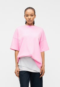 Νεαρός με πλεγμένα μαλλιά φορώντας φαρδιά ροζ κοντομάνικη μπλούζα πάνω από ένα στρωμένο λευκό πουκάμισο και σκούρο παντελόνι, στέκεται μπροστά σε ένα απλό φόντο.