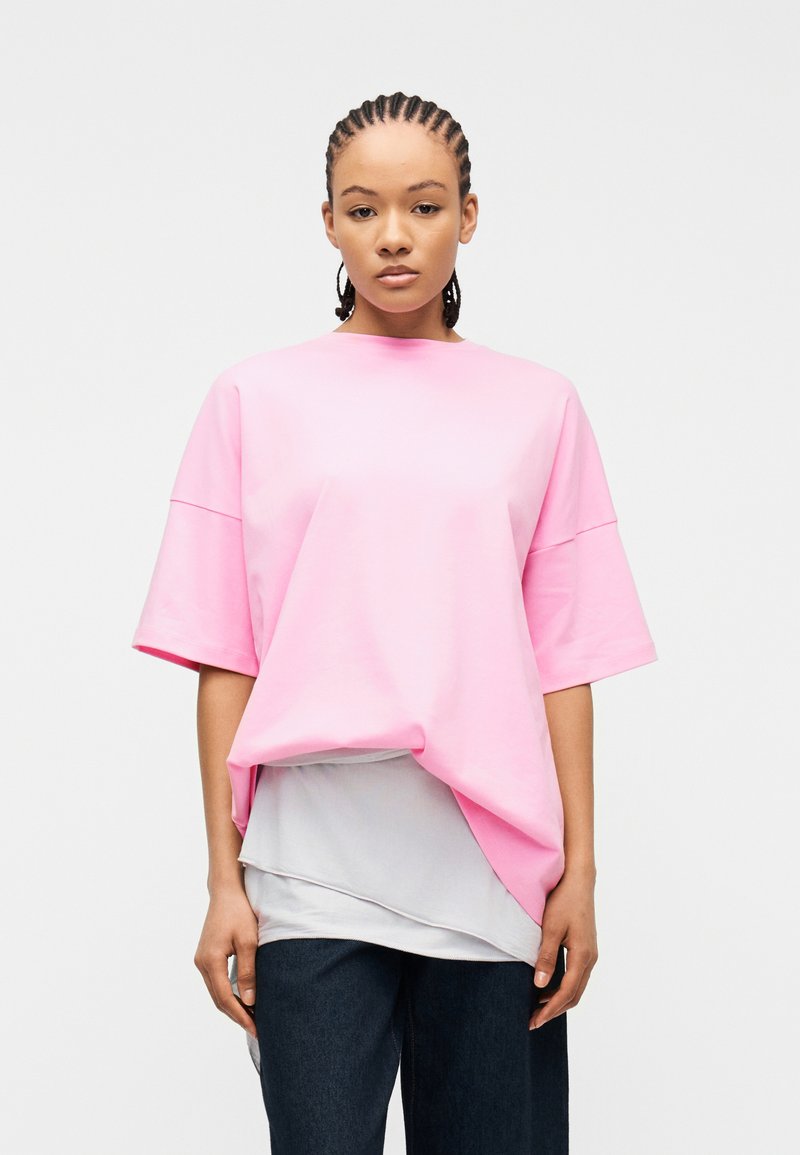 Νεαρός με πλεγμένα μαλλιά φορώντας φαρδιά ροζ κοντομάνικη μπλούζα πάνω από ένα στρωμένο λευκό πουκάμισο και σκούρο παντελόνι, στέκεται μπροστά σε ένα απλό φόντο.