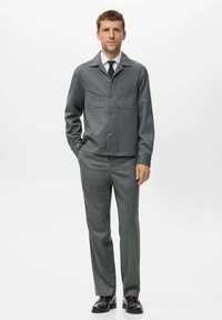 Traje gris a medida con una chaqueta abotonada que presenta bolsillos en el pecho, combinado con pantalones de pierna recta y zapatos de cuero negros.