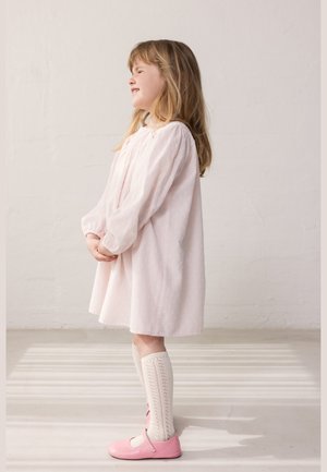 Jeune fille portant une robe rose clair, des chaussettes blanches jusqu'aux genoux et des chaussures roses, debout de profil, les mains jointes, souriante sur un fond neutre.