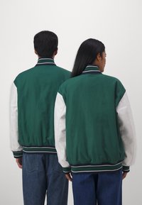 Casaco varsity verde com mangas brancas, gola e punhos canelados. Apresenta uma textura suave e riscas horizontais na gola e nos punhos.