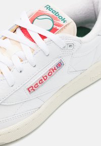Sapatilha de couro branca com superfície texturizada, logótipo Reebok vermelho e gráfico colorido de ténis na língua. Realçada com atacadores brancos.