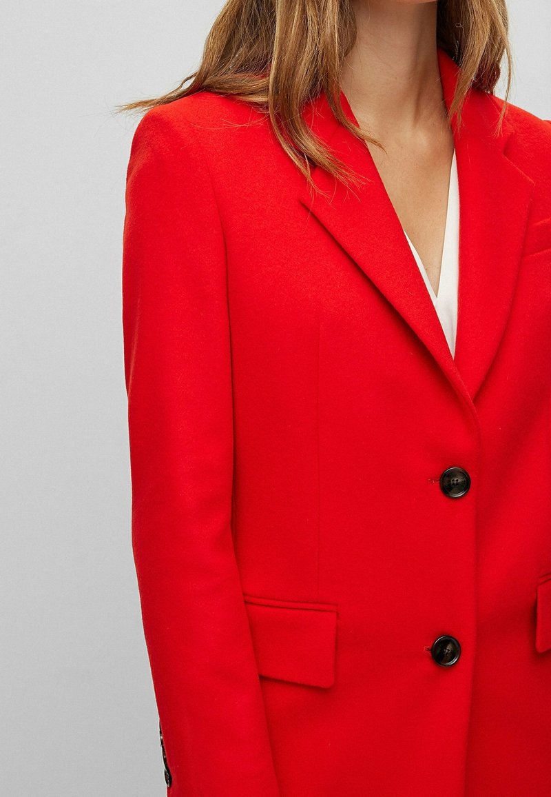 Femme portant un blazer rouge vif avec des boutons noirs sur un haut blanc, montrée de l'épaule jusqu'au milieu du torse sur un fond uni.