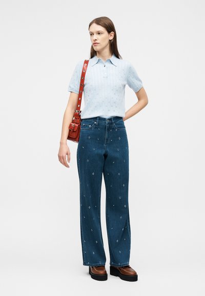 Γυναίκα που στέκεται φορώντας ένα ανοιχτό μπλε πουκάμισο με κοντά μανίκια, σκούρα μπλε κεντημένα τζιν, καφέ πλατφόρμες παπούτσια και κόκκινη τσάντα ώμου.