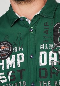 Chemise boutonnée verte avec un col intérieur blanc contrastant. Comprend des graphismes en texte audacieux, un patch rond et des accents de boutons foncés.