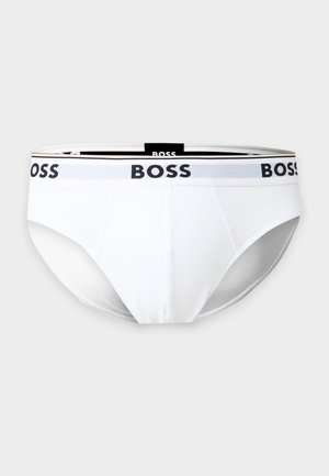 Homme athlétique debout pieds nus, portant un slip blanc avec le logo noir « BOSS » sur la ceinture, sur fond blanc uni.