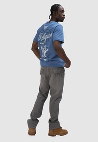 Camiseta de algodón de color azul claro con un diseño gráfico en la parte posterior, combinada con jeans de mezclilla grises y botas de trabajo marrones.