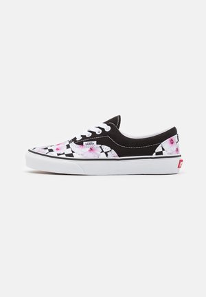 Sort-hvid Vans sneakers med skakbræt- og lyserødt blomstermønster, hvide snørebånd og hvid sål, set fra siden mod en ensfarvet baggrund.