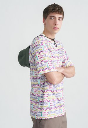 Noor mees heleda nahaga ja lokkis juustega, kandes värvikat mustrilist särki ning seljas rohelise ämbritüüpi mütsi, käed rüpes.