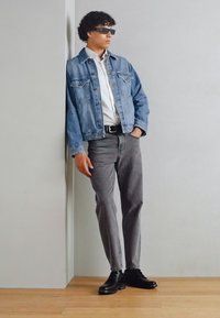 Jeansowa kurtka, biała koszula, szare spodnie i czarne buty. Model opiera się o ścianę, nosząc okulary przeciwsłoneczne. Swobodny strój z różnymi teksturami.
