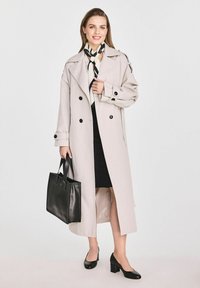 Jasno-beżowy trencz z podwójnymi guzikami, czarnymi akcentami, noszony na czarnej sukience. Czarna skórzana torba na ramię i buty na słupku.