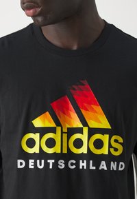 Czarny bawełniany t-shirt z dużym logo w kształcie trójkąta w gradientzie od czerwonego do żółtego oraz napisem "adidas DEUTSCHLAND" w białym i żółtym.