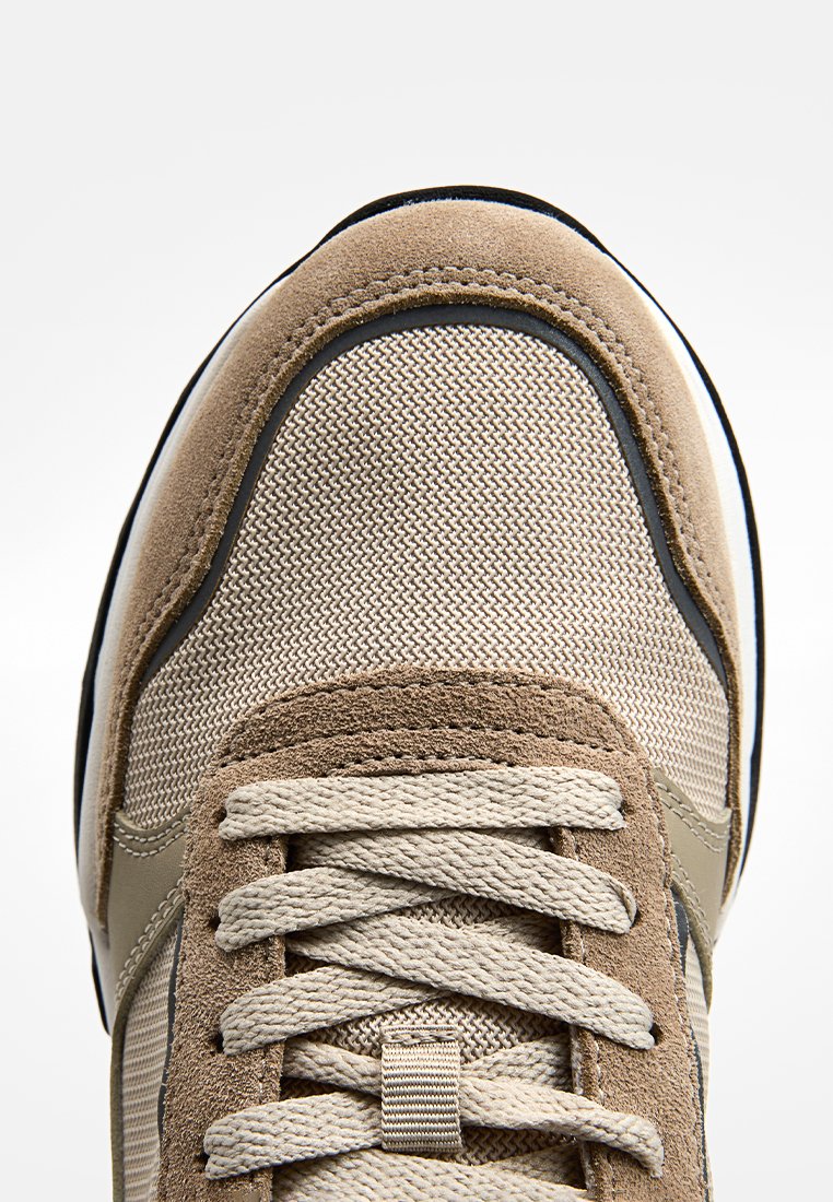Braune und beige Sneaker aus gemischten Materialien mit strukturiertem Finish, atmungsaktivem Mesh, Schnürsenkeln und einem schwarzen Akzent entlang der Sohle.