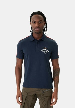 Uomo con capelli intrecciati indossa una polo blu navy con il logo "Aeronautica Militare 1923" e pantaloni cargo oliva, in piedi su sfondo semplice.