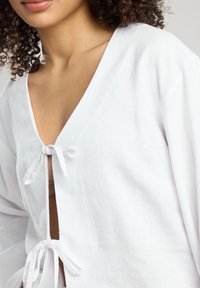 Blusa bianca in lino con scollatura a V profonda e chiusure annodate sul davanti. Presenta maniche lunghe e ampie e una finitura morbida e testurizzata.