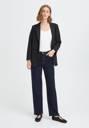 Donna in piedi con la mano in tasca, indossa un blazer nero, una maglietta bianca, jeans larghi scuri e scarpe basse nere su sfondo semplice.