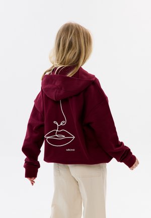 Osoba o blond włosach nosi bordową bluzę z kapturem z białym, ciągłym rysunkiem ust i nosa na plecach, w połączeniu z beżowymi spodniami.