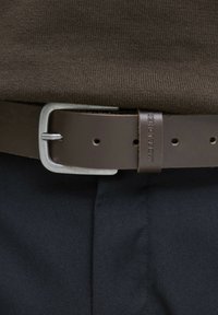 Brown leather belt with a silver rectangular buckle and engraved brand label, worn over dark fabric, showing texture and design details.