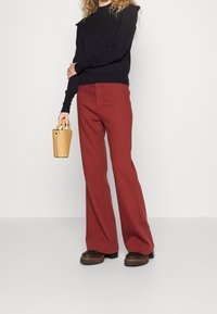 Osoba ubrana w czarną bluzkę z długim rękawem i szerokie spodnie w kolorze rdzy, trzymająca małą beżową torebkę, stojąca na białej podłodze.