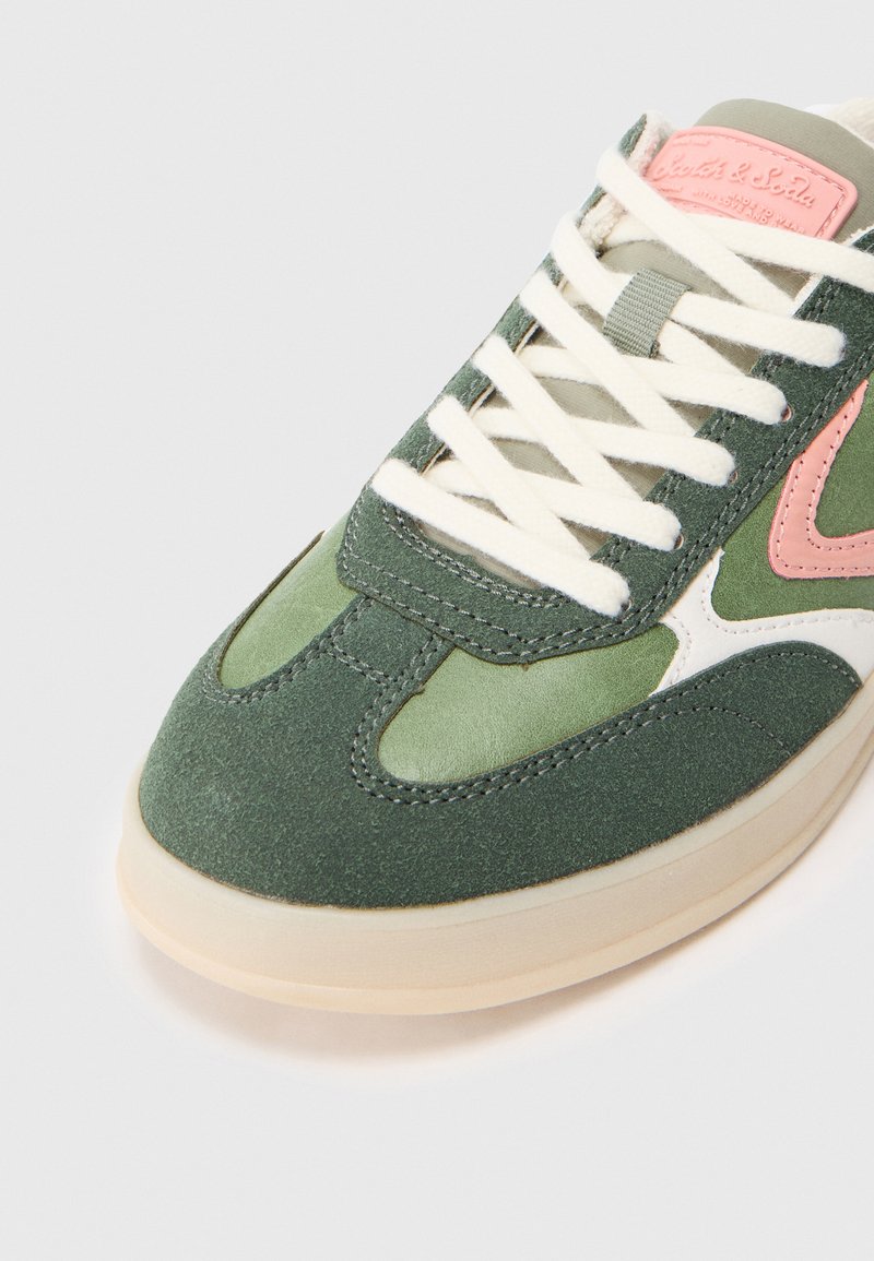 Groene en beige sneaker met een gestructureerde suède bovenkant, roze accenten en witte veters. De vorm heeft een ronde neus en een stevige zool.