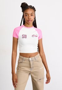 Camiseta corta blanca con mangas raglán rosas, que presenta bordado de "59" y el logo de "ellesse". Combinada con jeans rotos de color beige claro.