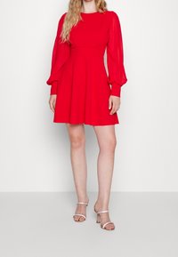 Punainen mekko, jossa on pitkät, tilavat hihat ja istuva yläosa, joka levenee hameeksi. Yhdistetty hopeisiin korkokenkiin. Silkkinen kangaslaatu.