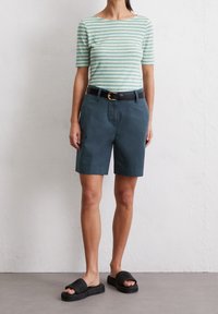 Mujer vistiendo una blusa de rayas verde y blanca de mangas cortas, pantalones cortos oscuros de color teal con cinturón y sandalias negras de tiras, de pie contra una pared lisa.
