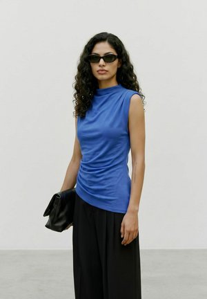 Femme aux cheveux bouclés portant des lunettes de soleil noires, un haut bleu sans manches, un pantalon large noir et tenant une pochette noire.
