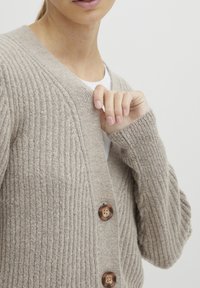 Beige ribbestrikket cardigan med v-hals, lange ermer og tre store brune knapper foran. Myk tekstur og avslappet passform.