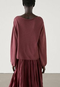 Maglione lavorato a maglia color bordeaux con maniche a palloncino, vestibilità oversize e design a schiena aperta, abbinato a una lunga gonna fluida e plissettata dello stesso colore.