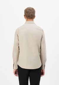 Man met kort krullend haar, gekleed in een beige lange mouwen overhemd met knopen en zwarte broek, staand met zijn rug naar de camera tegen een witte achtergrond.