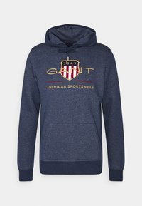 Sudadera azul oscuro con bolsillo canguro, con un logo de escudo y "GANT" en bordado dorado y rojo. Tejido de mezcla de algodón.