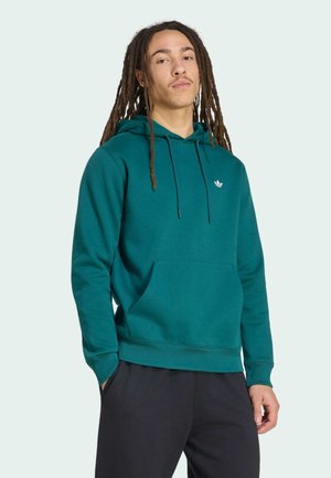 Młody mężczyzna z długimi dredami, ubrany w turkusową bluzę adidas z kapturem i czarne spodnie, stoi z jedną ręką w kieszeni na jasnym tle.