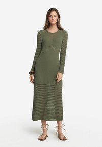 Olivgrön stickad klänning med långa ärmar, med öppen arbetsmönster, en figurnära bodice och en flödande botten. Matchad med sandaler med rem. 