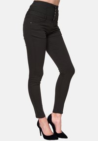 Jeans skinny neri a vita alta con una texture liscia, chiusura con cinque bottoni e dettagli di cuciture sottili. Indossati con tacchi neri alti.