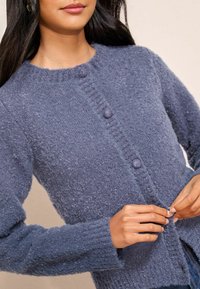 Moteris sagsto mygtukus ant tekstūruoto, ilgomis rankovėmis, mėlyno mezginio kardigano su apvalia iškirpte.