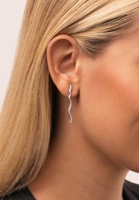 Silberne, schlangenförmige Ohrringe mit einer glatten Oberfläche. Leichtes, hängendes Design betont das Ohr. Minimalistischer Stil, passend zu verschiedenen Outfits.